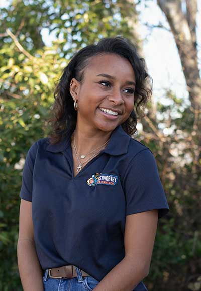 Smiling female Marketing & Operations Administrator from Trustworthy Electric wearing a branded polo shirt, outdoors with greenery in the background.