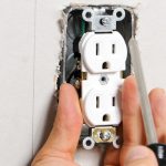 Hand holding screwdriver installing tamper-resistant electrical outlet in wall, emphasizing safety features for homes with children.